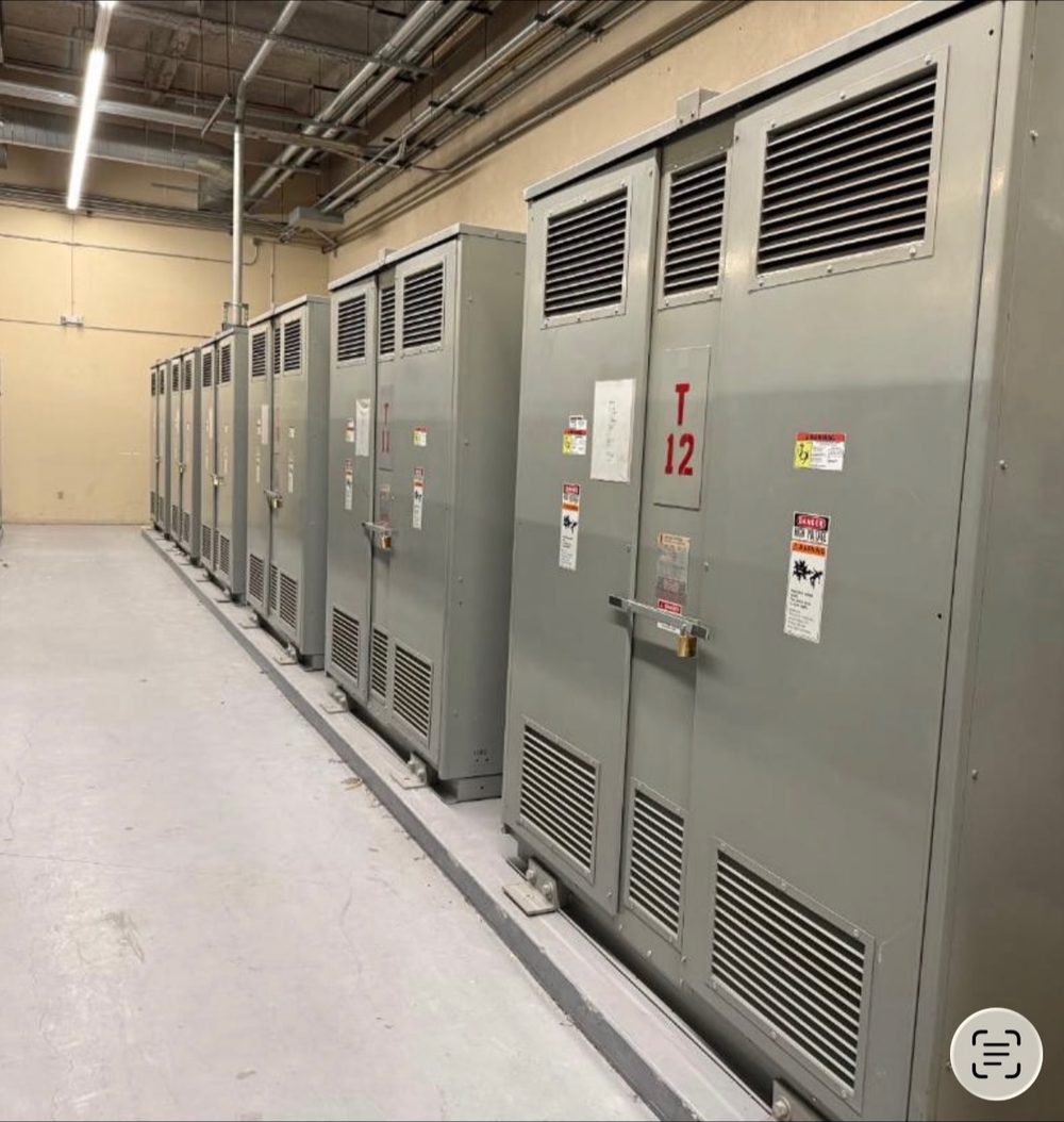 Long row of grey medium voltage switchgear and unit substation sections with louvered ventilation panels inside an electrical room, tagged and ready for removal